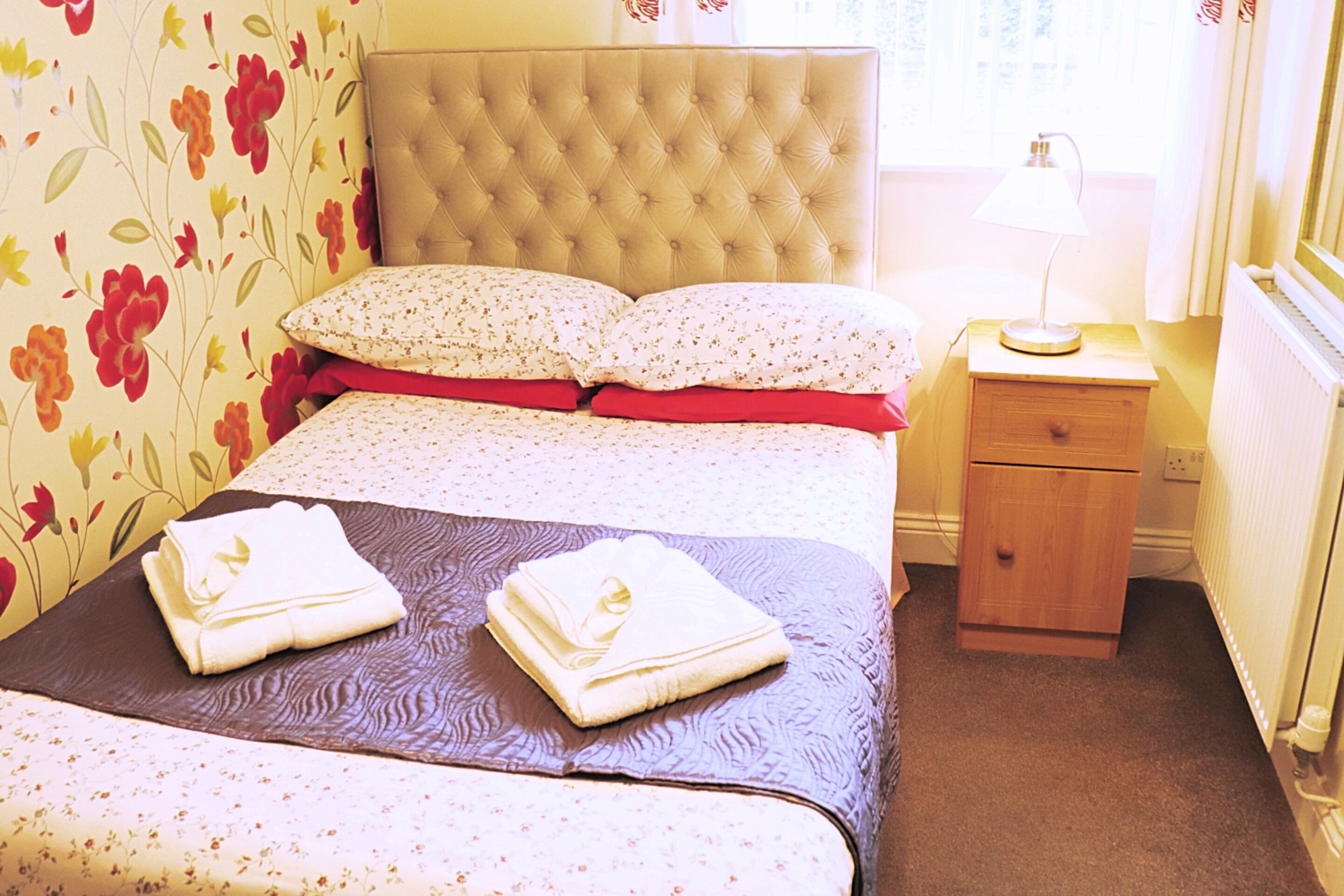 Una cama bien hecha con un cabecero estampado con motivos florales, una mesita de noche con una lámpara y un papel tapiz floral en la pared.