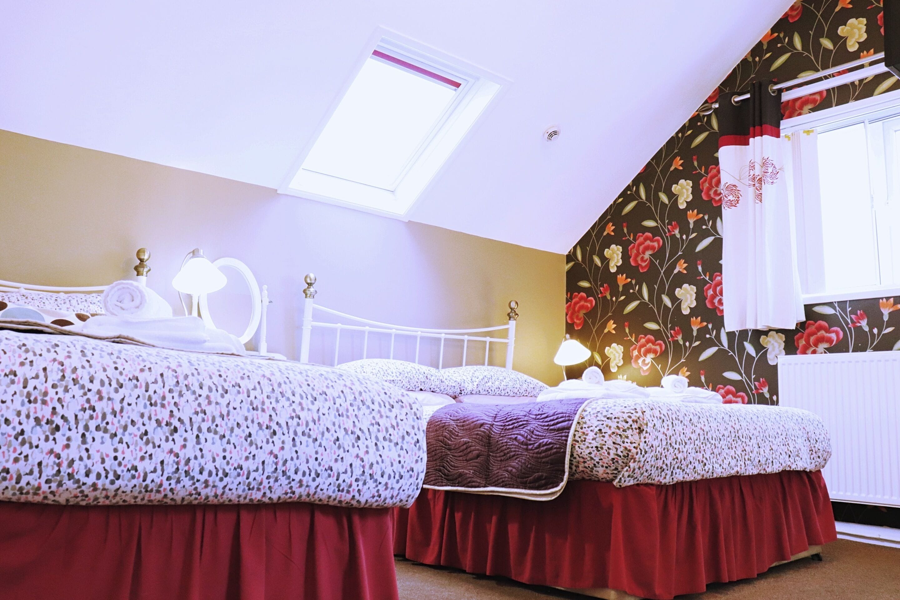 A bedroom with two beds, a skylight, floral wallpaper, and a radiator.