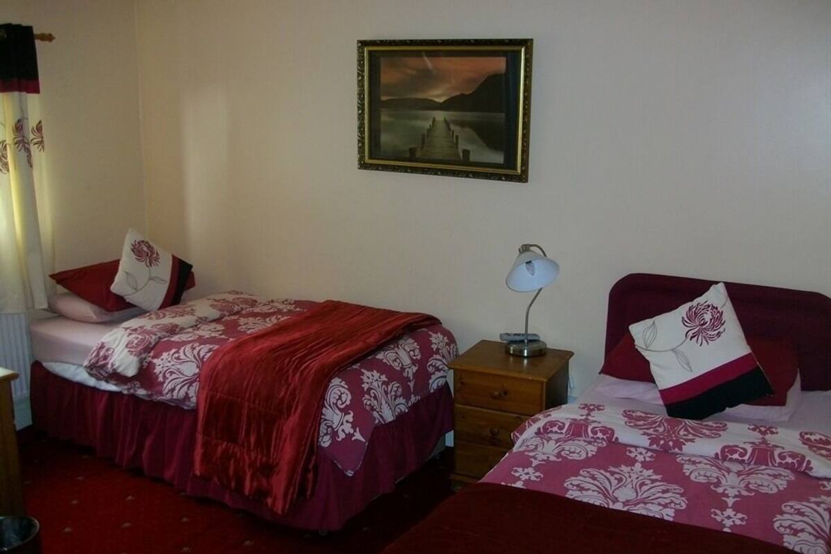 A room with two beds, each with a red and white patterned bedspread, a nightstand with a lamp, and a framed picture on the wall.