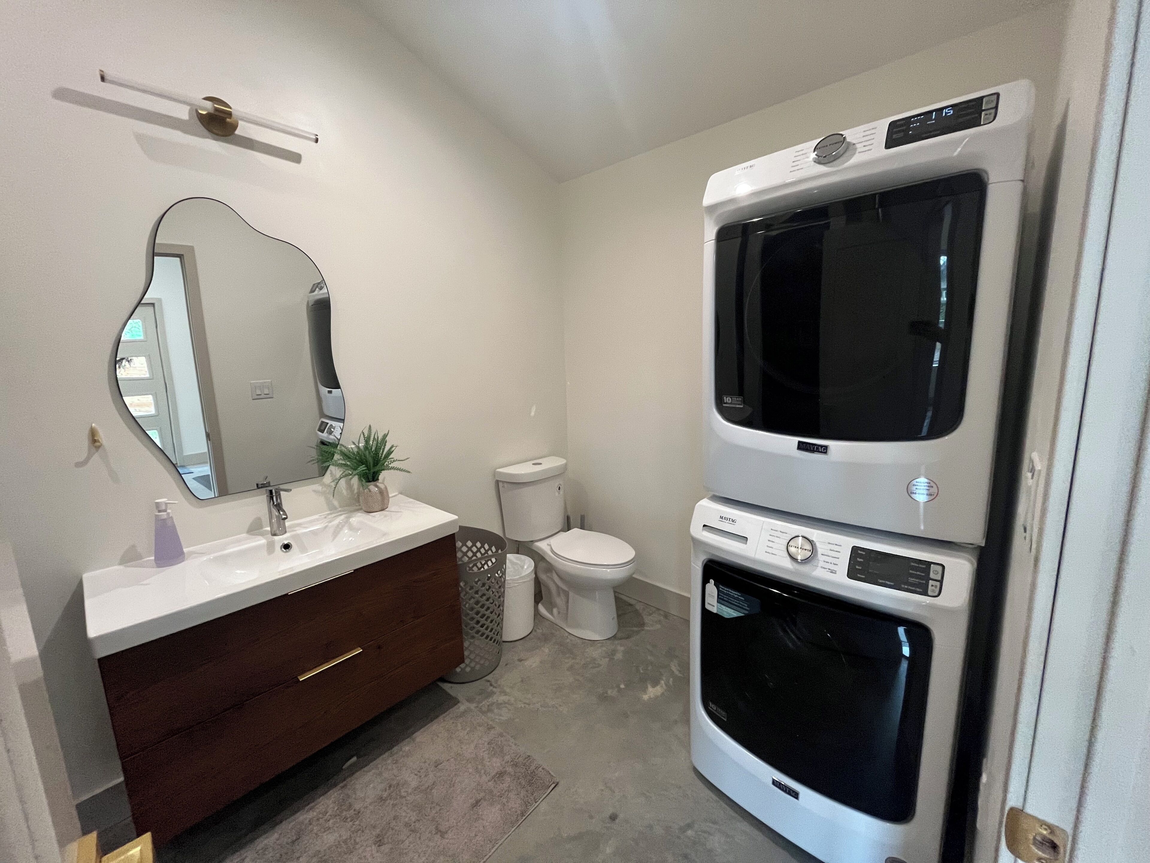 Une salle de bain avec un lavabo blanc, un miroir, des toilettes et des appareils électroménagers superposés, notamment un lave-linge et un sèche-linge.