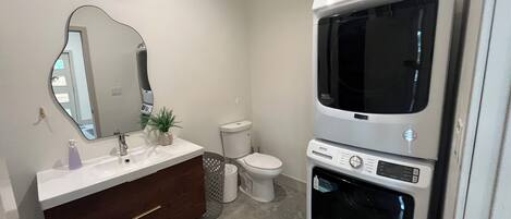 Une salle de bain avec un lavabo blanc, un miroir, des toilettes et des appareils électroménagers superposés, notamment un lave-linge et un sèche-linge.