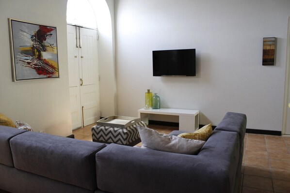 A living room with a grey sofa, a flat-screen TV mounted on the wall, a white cabinet, and a door with a handle.