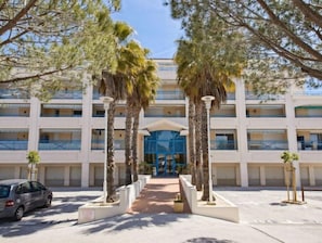 Un hotel a più piani con un ingresso centrale, palme e un'auto parcheggiata di fronte.