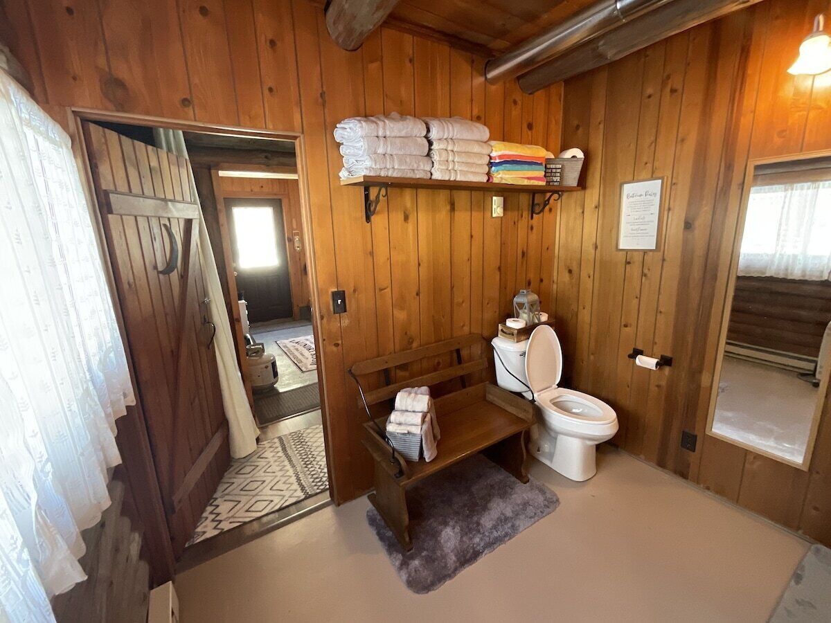 Un baño con paneles de madera, que incluye un inodoro, un banco y una estantería con toallas dobladas.