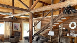 Interior espacioso de una cabaña de troncos con techos altos, una escalera y una zona de estar acogedora con varios sofás y sillones.