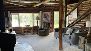 Interior rústico de una cabaña con chimenea de piedra, un sofá, un sillón y un ventanal con vistas a un lago.