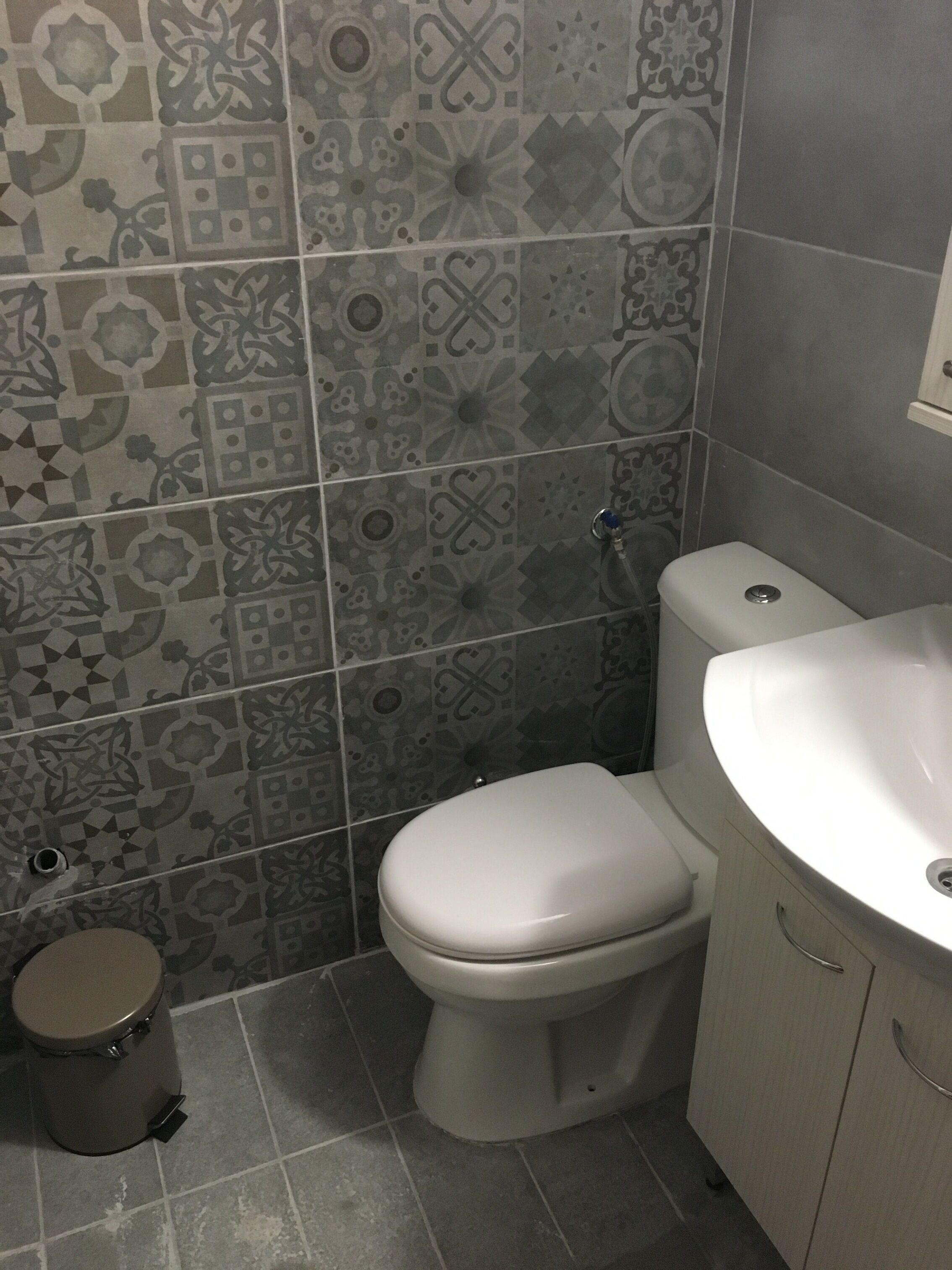 A bathroom with a toilet, sink, and trash can. The walls are tiled with a patterned design.