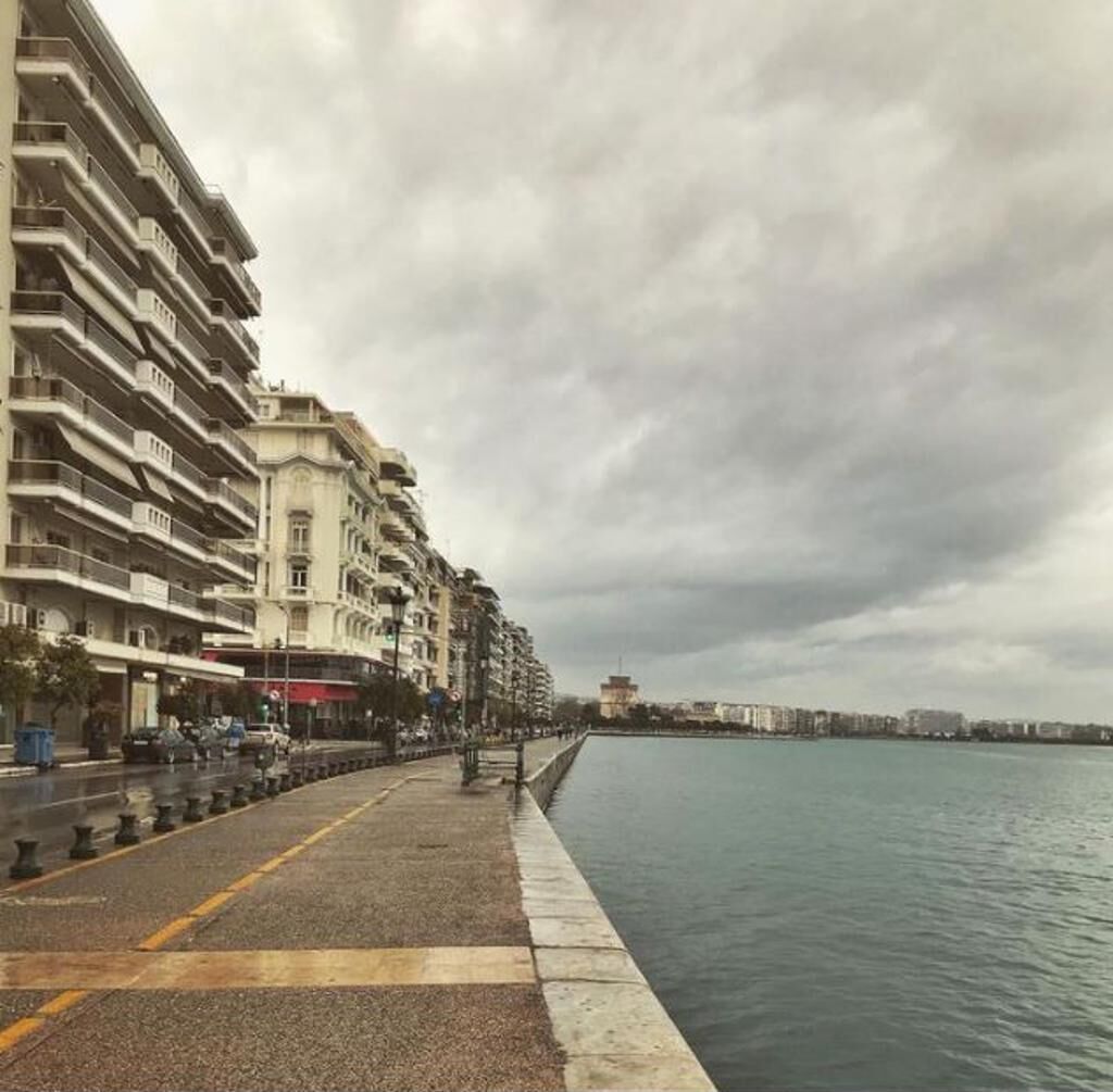 A waterfront promenade with multi-story buildings, a calm body of water, and a cloudy sky.