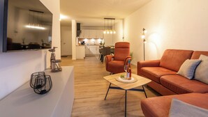 A modern living room with a sofa, armchair, and coffee table. A kitchen is visible in the background.