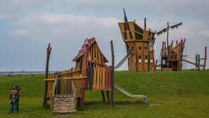A playground with a slide and climbing structures, set on a grassy hill with a view of the sea in the background.