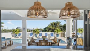 Un espacio moderno para comer al aire libre con vista a la piscina, que incluye una mesa y sillas de madera, y dos lámparas colgantes de mimbre.