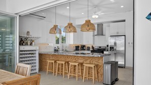 Una cocina moderna con armarios blancos, una isla de madera con taburetes de bar, lámparas colgantes de mimbre y un refrigerador de acero inoxidable.