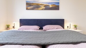 A bedroom with a large bed, a dark headboard, two bedside tables with lamps, and a wall-mounted painting of a beach scene. - Dorumer Sielhuus 13 mit Sauna und Balkon (Dorum-Neufeld)
