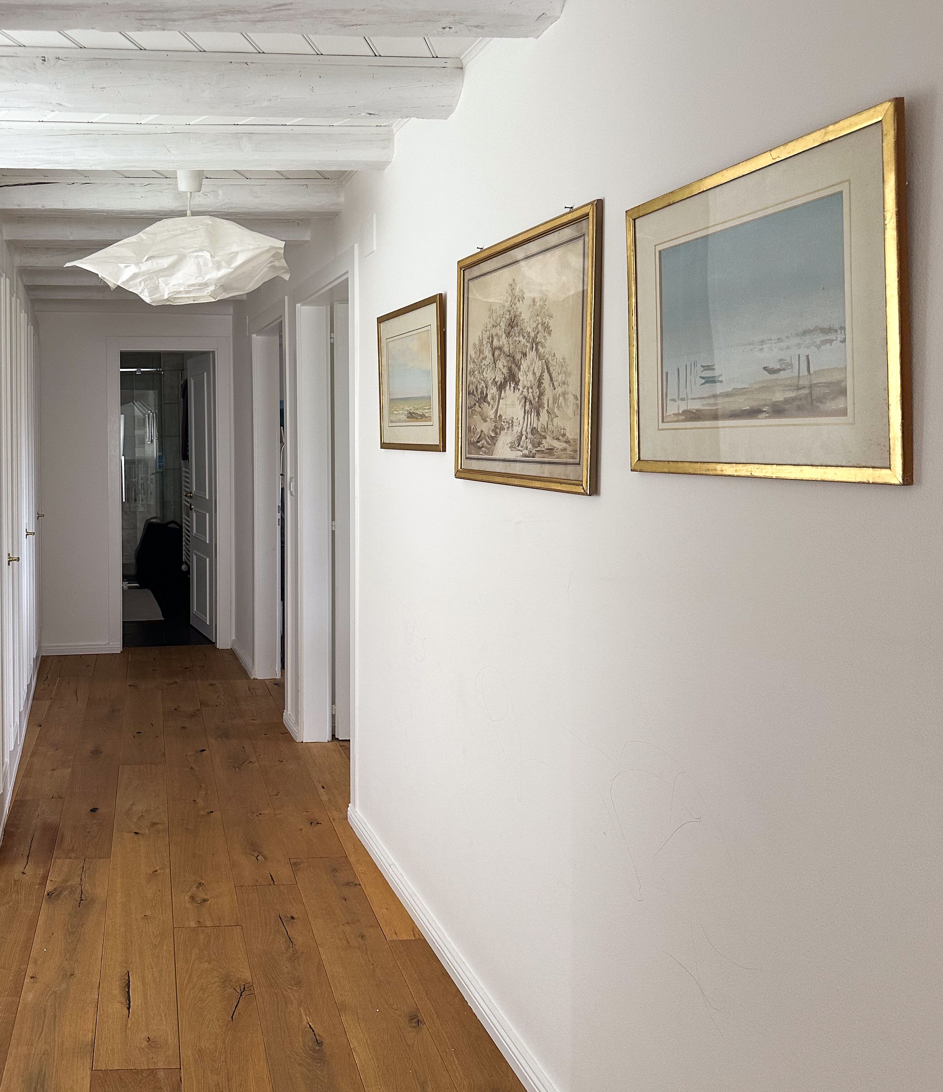 Un couloir doté d’un plancher en bois, de murs blancs et de trois œuvres d’art encadrées.
