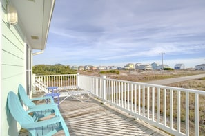 A wooden deck with a white railing, two blue Adirondack chairs, and a view of a beach and houses in the distance.