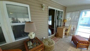 A sunlit room with a wicker chair, a cushioned ottoman, a small table with a lamp, and a sliding glass door leading to another room.