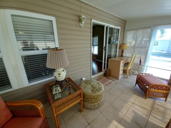A sunlit room with a wicker chair, a cushioned ottoman, a small table with a lamp, and a sliding glass door leading to another room.