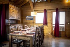 A wooden dining room with a table set for a meal, wooden chairs, a chandelier, and a view of the outside through large windows.