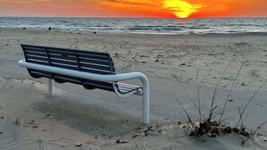 Eine Bank an einem sandigen Strand mit dem Ozean und einem Sonnenuntergang im Hintergrund.