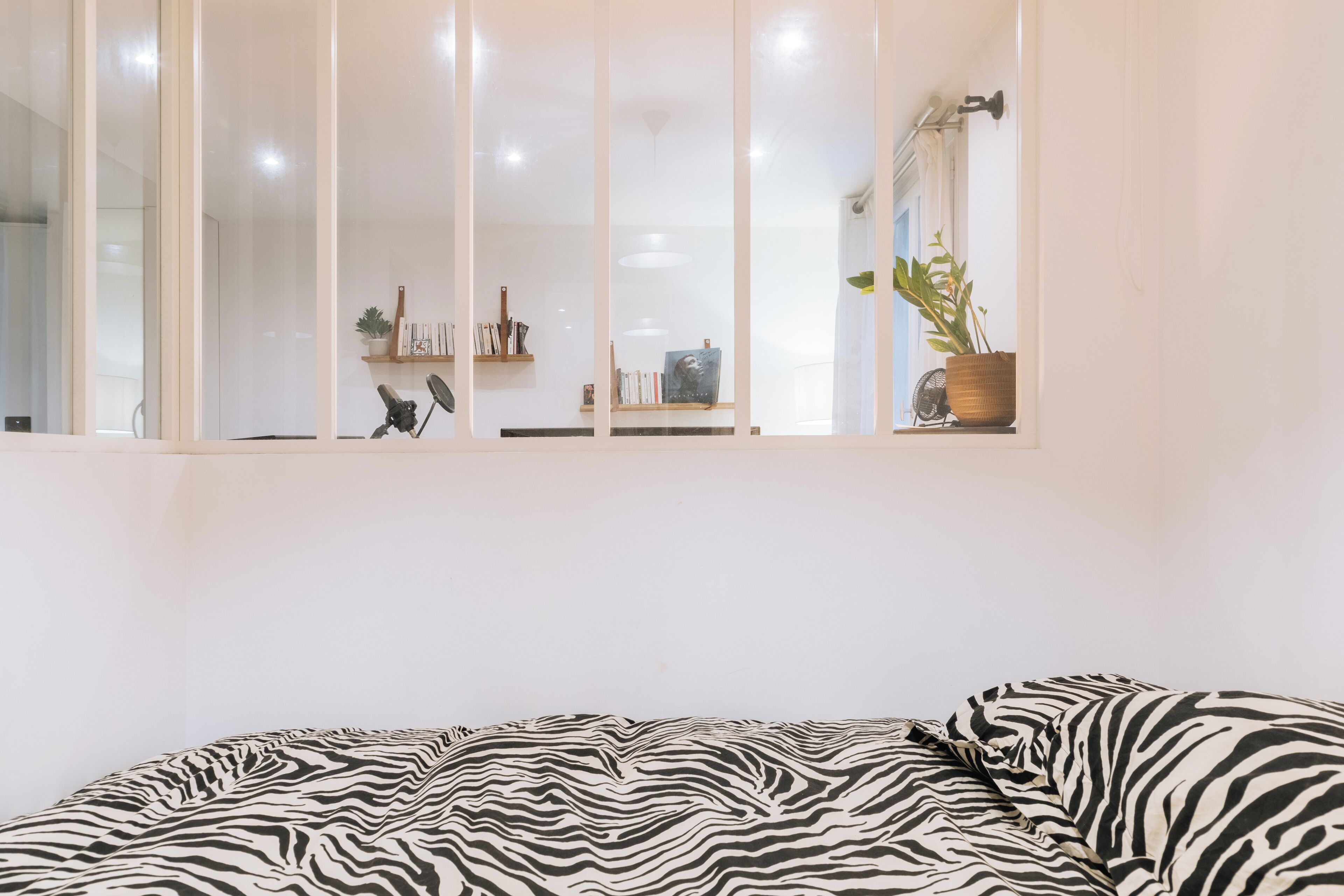 A bedroom with a zebra-patterned bedspread, a window with blinds, a potted plant, and a shelf with books and decorative items.