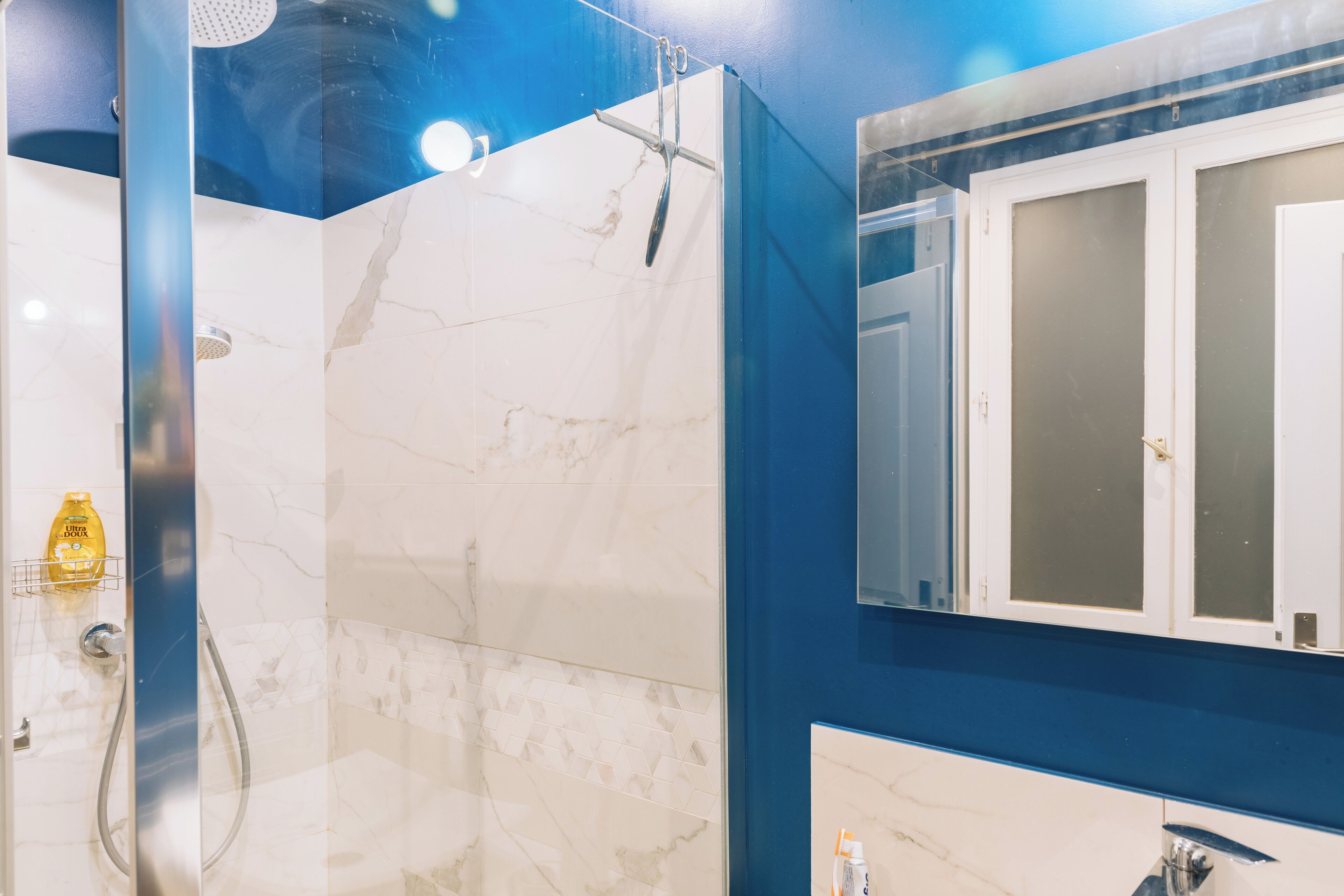 A bathroom with a glass-enclosed shower, marble tiles, a blue accent wall, and a mirror with frosted glass panels.