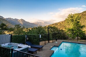A pool with lounge chairs and a hot tub, surrounded by a fence and trees, with mountains in the background.