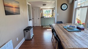 Un coin repas avec une table en bois, des chaises, une horloge et une vue sur la cuisine et le jardin.