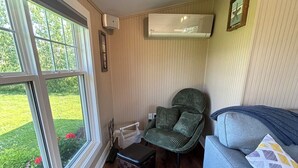 Une pièce avec un plancher en bois, un canapé gris orné de coussins à motifs, un fauteuil vert, une petite table en bois et une fenêtre donnant sur un jardin.