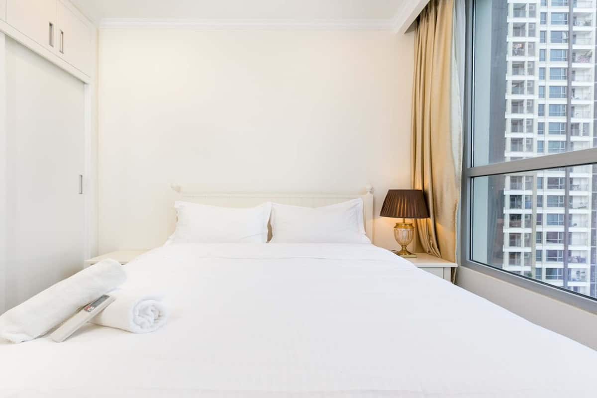 Quarto de hotel com uma cama grande, uma mesinha de cabeceira com abajur e uma janela com vista para a cidade.