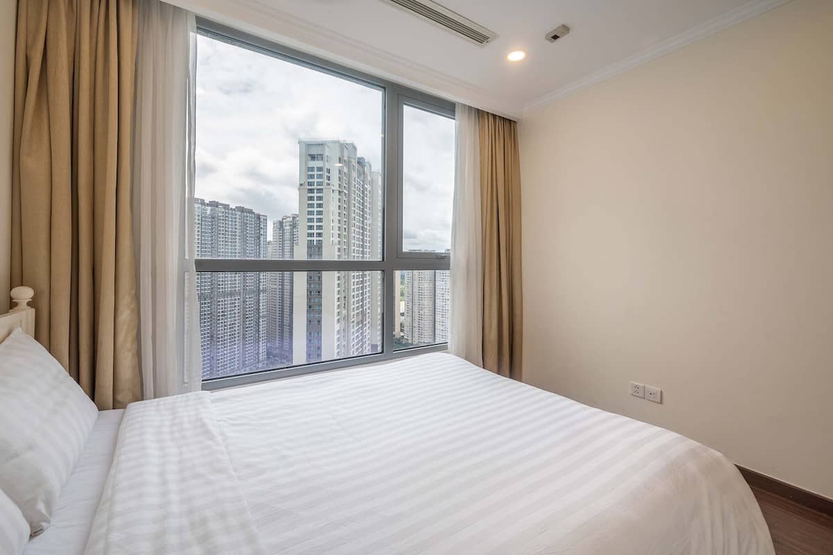 Quarto de hotel com uma janela grande, uma cama com roupa de cama branca e vista para edifícios altos.