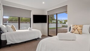 A modern bedroom with two beds, a flat-screen TV, and large sliding glass doors leading to a backyard. - Waterfront Oasis - Omokoroa Holiday Home (Omokoroa)