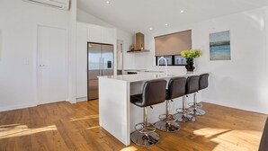 A modern kitchen with a white island and bar stools, a stainless steel refrigerator, and a large window with a view. - Waterfront Oasis - Omokoroa Holiday Home (Omokoroa)