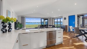 A modern kitchen with a large island, a dining area with a view of the water, and a living space with a blue door. - Waterfront Oasis - Omokoroa Holiday Home (Omokoroa)