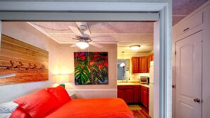 Ein Schlafzimmer mit einem großen Bett, einem Deckenventilator, einem Gemälde und Blick in eine Küche.
