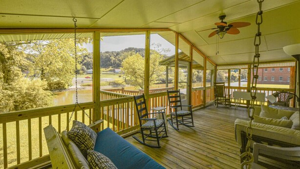 A covered porch with a wooden deck, seating area with a sofa and chairs, a ceiling fan, and a view of a river and trees. - Toccoa River House- Toccoa River Front-2br/2ba-wifi-walk to town-Free Activities (McCaysville)