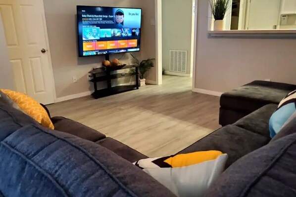 Un salon avec un téléviseur à écran plat fixé au mur, un canapé de couleur sombre agrémenté d’un coussin jaune et une petite table sur laquelle est posée une plante.