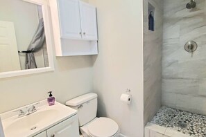 Une salle de bain avec un lavabo blanc, des toilettes, une douche avec un mur en marbre et un miroir au-dessus du lavabo.