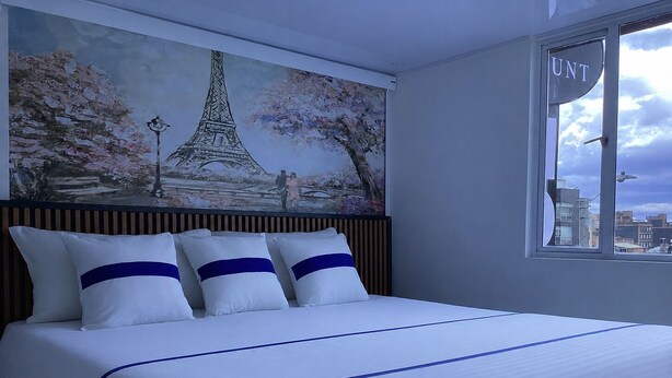 A hotel room with a bed, a painting of the Eiffel Tower, and a window with the 'UNT' sign. - Paramount Hotel (Bogotá)