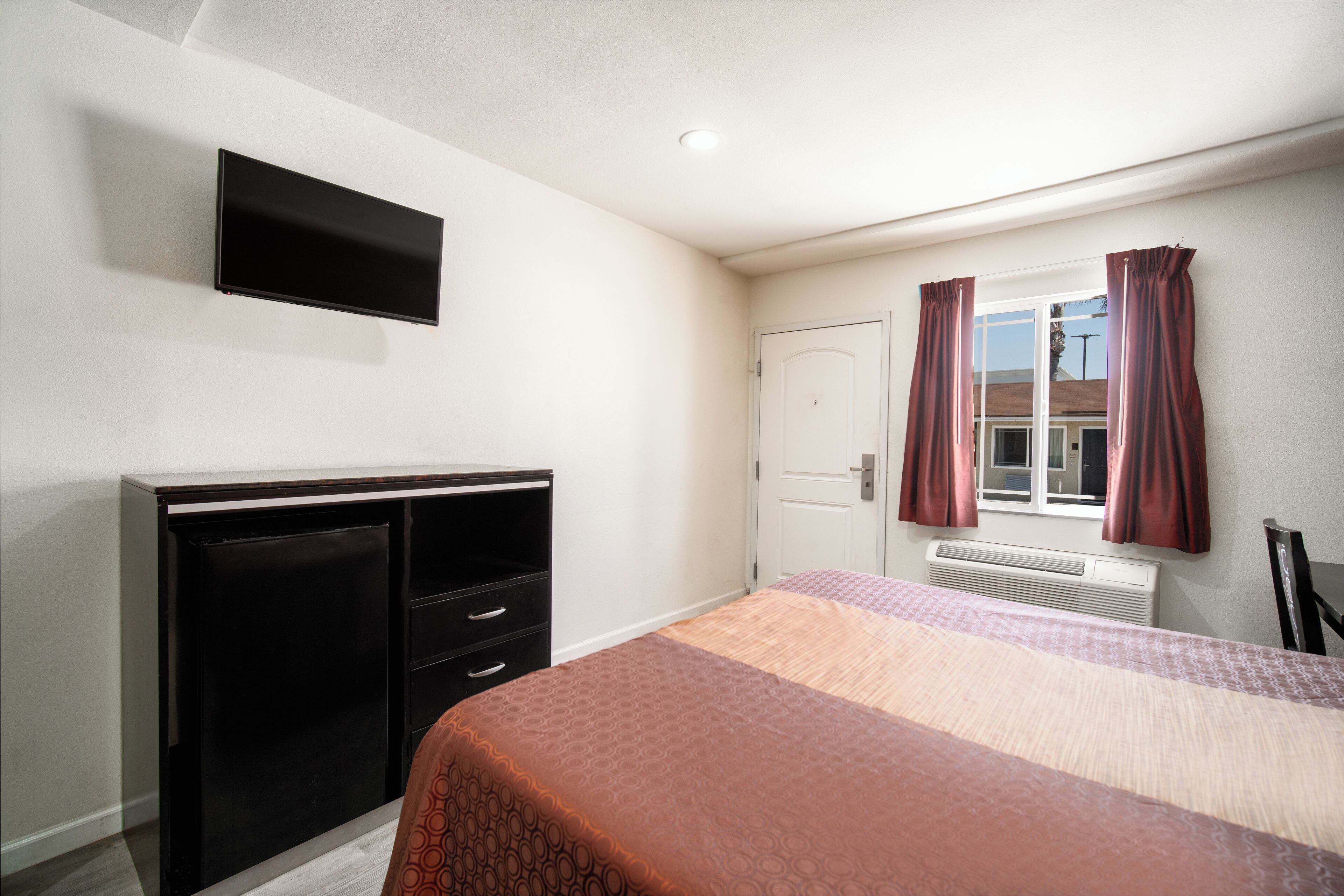 Un dormitorio con cama, cómoda, televisor montado en la pared y ventana con cortinas.