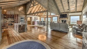 A spacious open-plan living area with a high ceiling, wooden beams, a kitchen with rustic cabinetry, and a seating area with a fireplace. - Heated pool & hot tub at huge mountain retreat

 (Cherry Log)