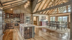 A spacious kitchen with wooden beams, a dining area with a large table and chairs, and a view of the outdoors through large windows. - Heated pool & hot tub at huge mountain retreat

 (Cherry Log)