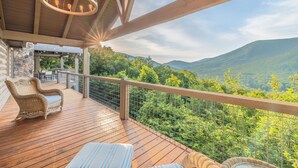 A wooden deck with wicker furniture, a stone wall, and a view of a forested mountain range. - Heated pool & hot tub at huge mountain retreat

 (Cherry Log)