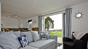 A modern living room with a grey sofa, a dark armchair, and a view of the outdoors through large windows. - Wheal Tor Lodge (Helston)