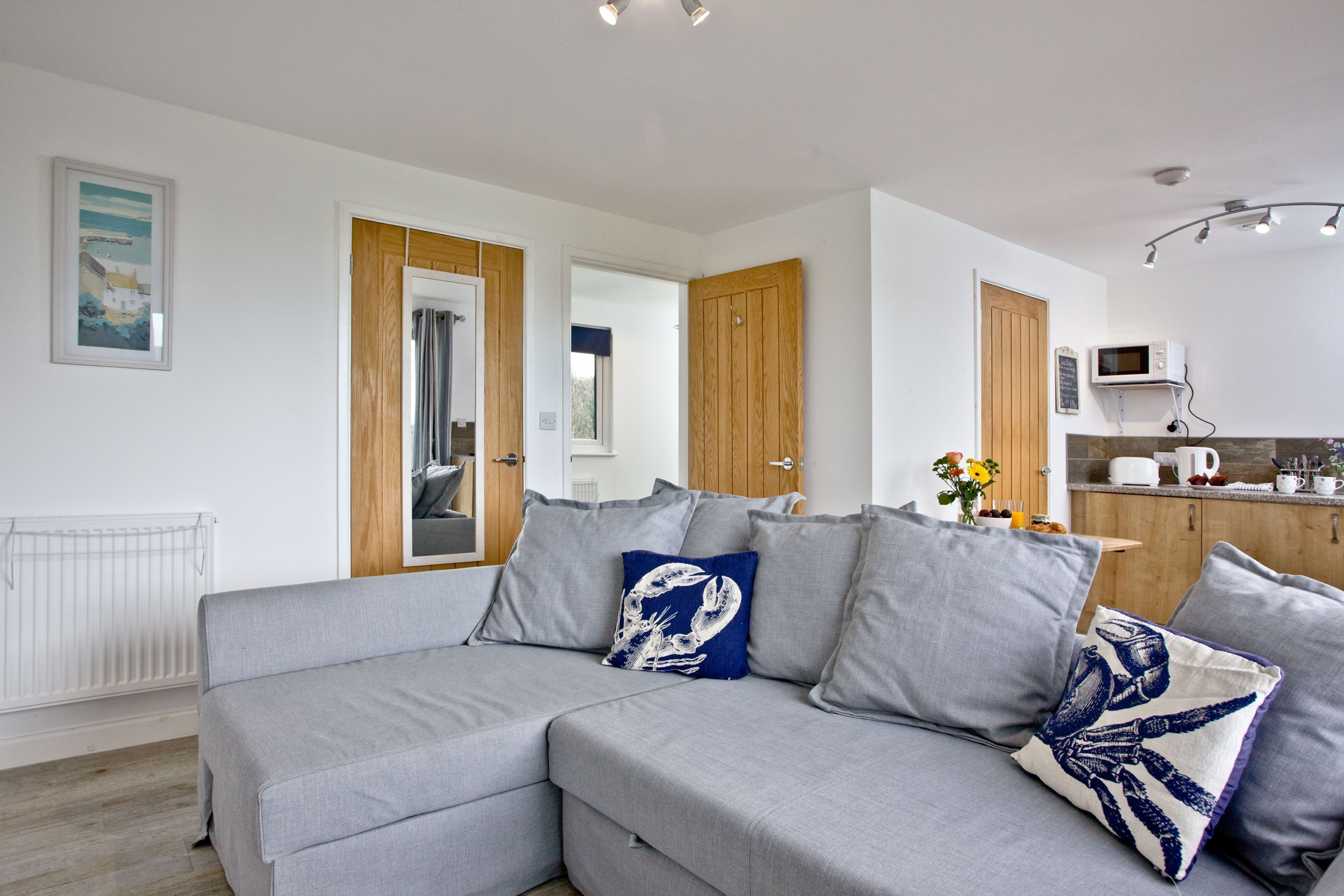 A modern living room with a grey sectional sofa, wooden doors, a white radiator, and a kitchen area with a microwave.