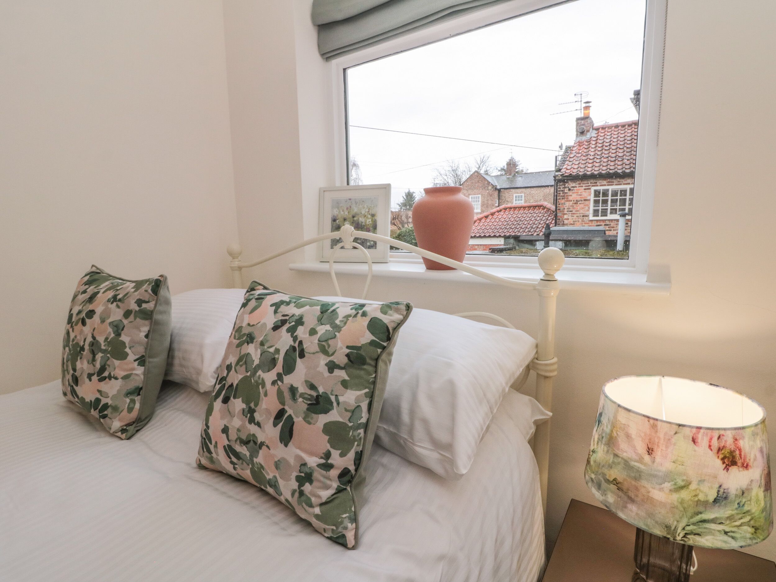 A bedroom with a bed, two pillows, a bedside lamp, a window, and a vase on the windowsill.
