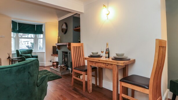 Un coin repas accueillant avec une table en bois, deux chaises et un fauteuil vert. On aperçoit une cheminée au fond, et il y a une fenêtre donnant sur l’extérieur.