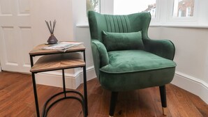 Un fauteuil vert placé à côté d’une table d’appoint en bois, sur laquelle se trouvent un magazine et un diffuseur de parfum en roseau.
