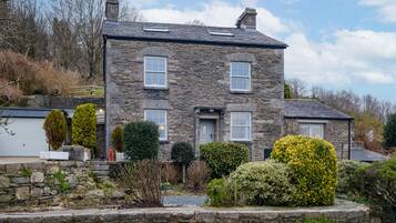 A stone house with a symmetrical facade, two visible windows, and a well-maintained garden with various shrubs and a stone wall.