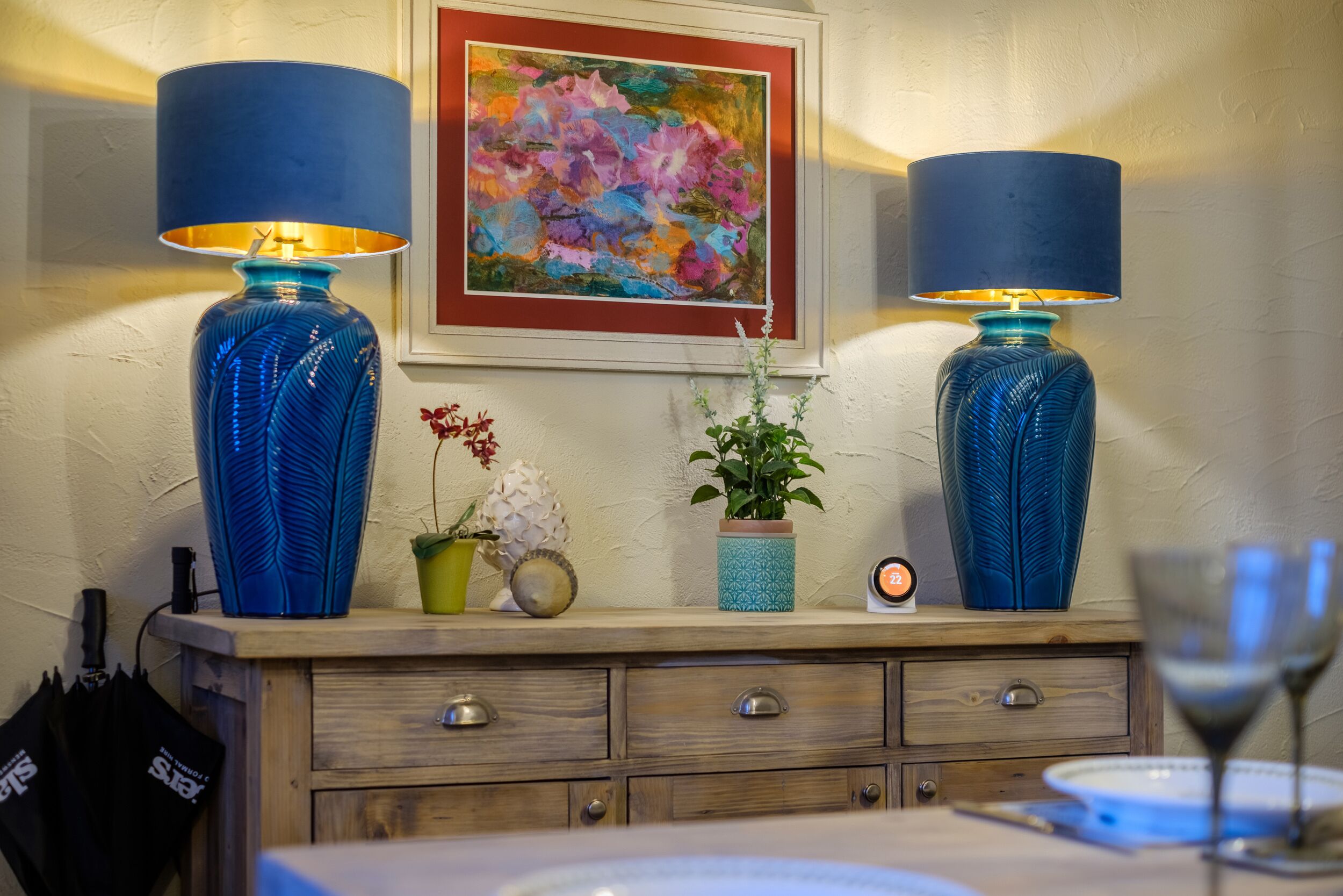A room with two blue lamps, a framed painting, a potted plant, a clock, and a wine glass on a table.