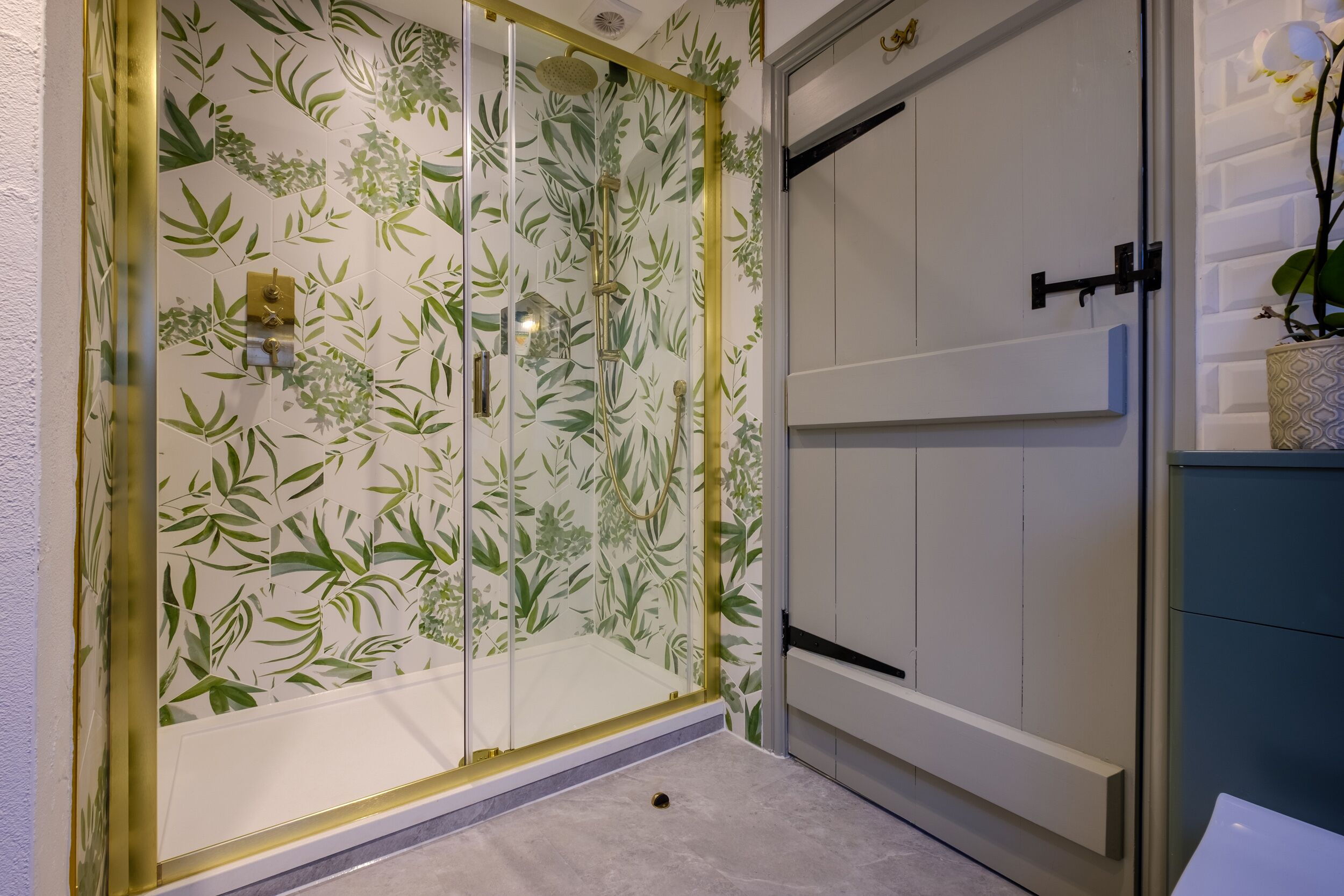 A bathroom with a shower enclosure featuring a glass door with a green leaf pattern, a white sink, and a blue cabinet.
